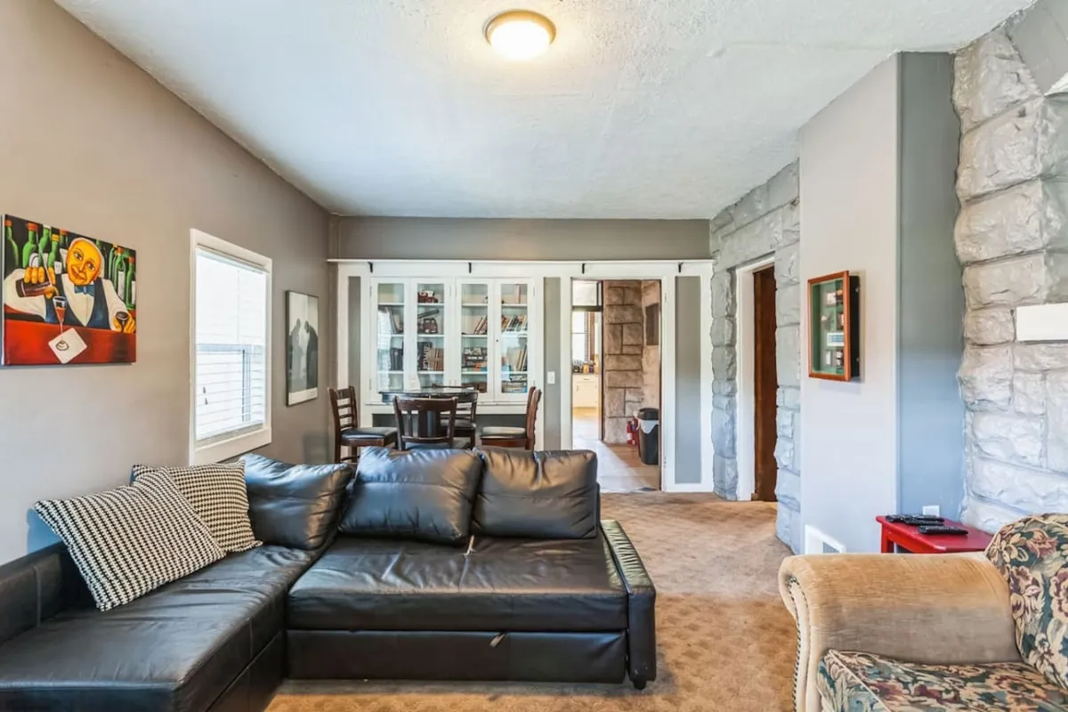 A cozy and welcoming living and dining area with a black leather sectional sofa, a large wooden dining table, and colorful artwork on the wall. This open space is designed for entertaining guests, family dinners, or informal gatherings, making it suitable for unwinding after productive sessions.