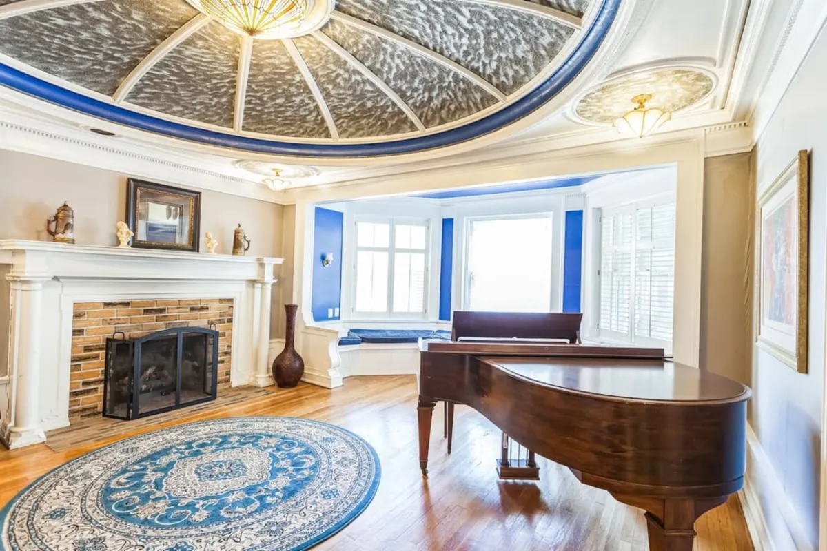 A sophisticated interior room featuring a grand piano, ornate ceiling, fireplace, and a cozy sitting area by a window. The space exudes warmth and refinement—perfect for relaxing after a day spent discovering local culture.