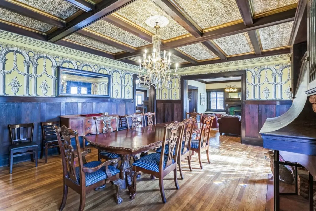 An opulent dining room with a long wooden table, ornate ceiling, chandelier, and decorative wall panels. The space suggests lavish dinners, family celebrations, and memorable events in a setting full of period detail.
