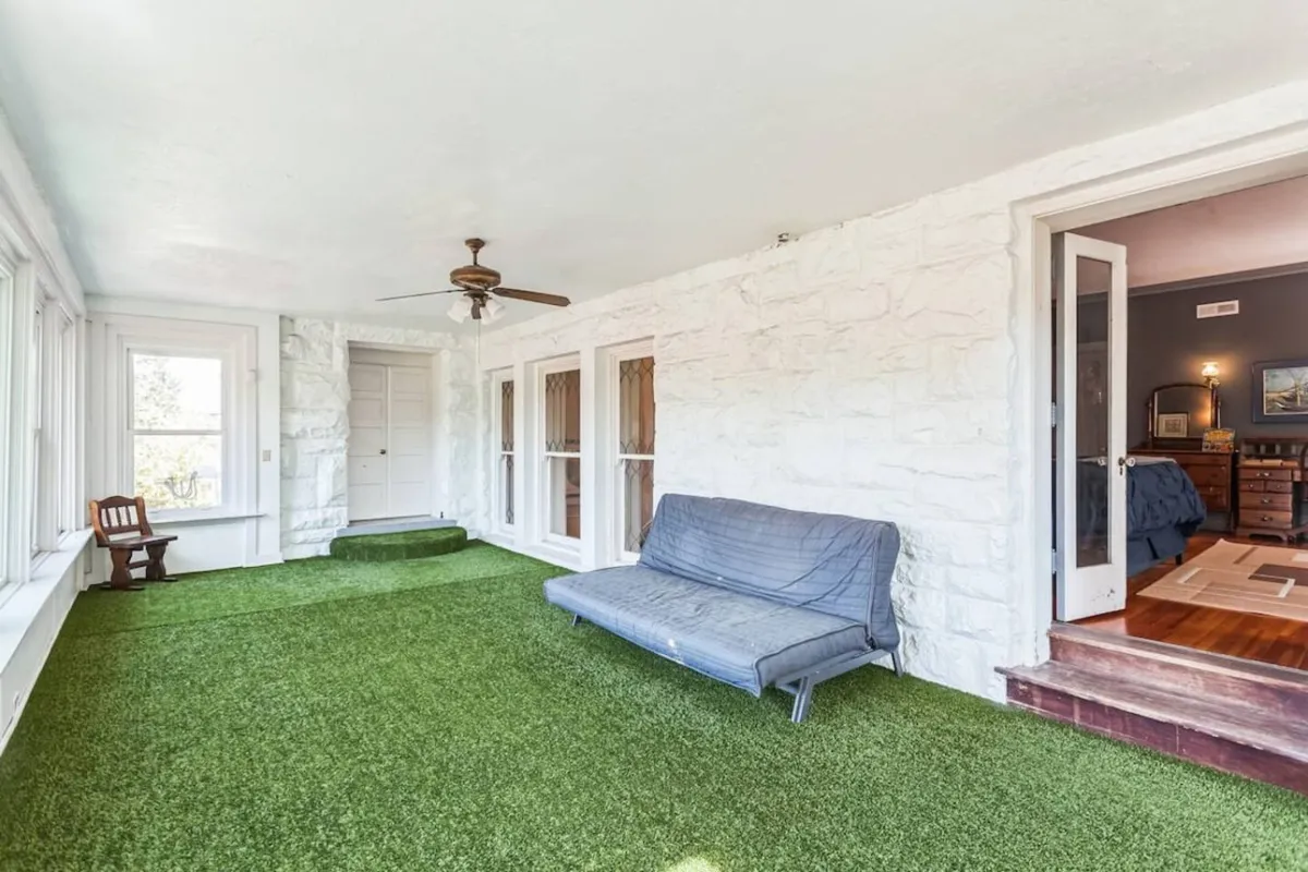 A unique indoor putting green space with green artificial grass flooring, a comfortable sofa, large windows allowing ample natural light, and a ceiling fan. The area feels casual, whimsical, and inviting—a playful spot that doubles as an extra sleeping area thanks to the queen futon mentioned in the section.