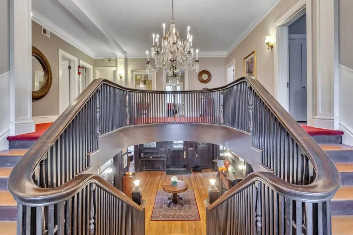 The image showcases the home's grand entryway, featuring an impressive staircase with intricate woodwork, a sparkling chandelier casting a warm glow, an ornate decorative mirror, a richly patterned area rug, and a cozy sitting area—perfect for welcoming guests. The atmosphere blends historical elegance with a sense of comfort, highlighting the home’s luxurious yet inviting character.