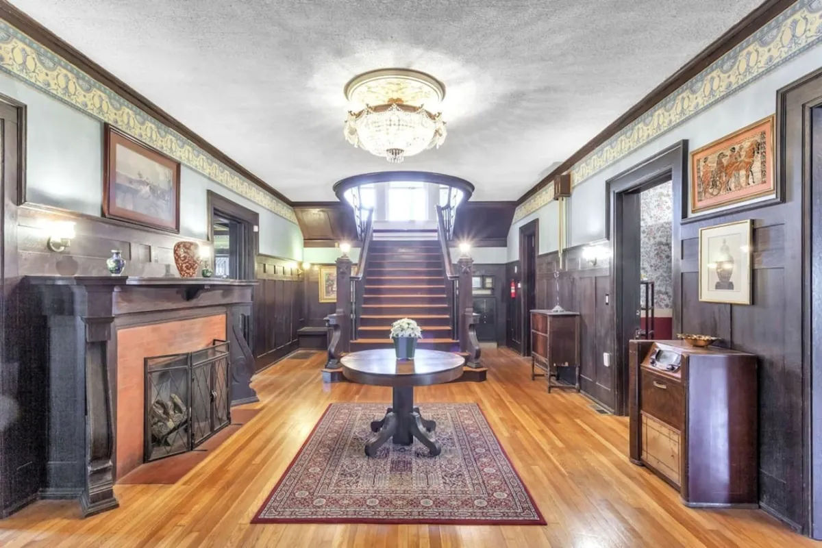 An elegant entrance area with a grand staircase, fireplace, round table, and chandelier, setting a welcoming tone for arriving guests and marking the start (or end) of an evening out.