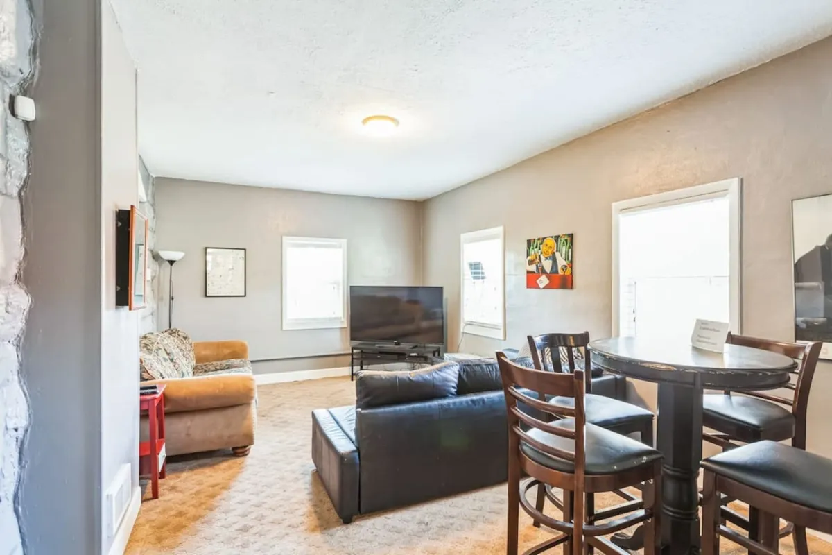 A flexible living area featuring a sofa, dining table, television, and prominent artwork. The relaxed setup hints at a TV room ideal for movie nights, socializing, or overflow sleeping, as well as casual gatherings.