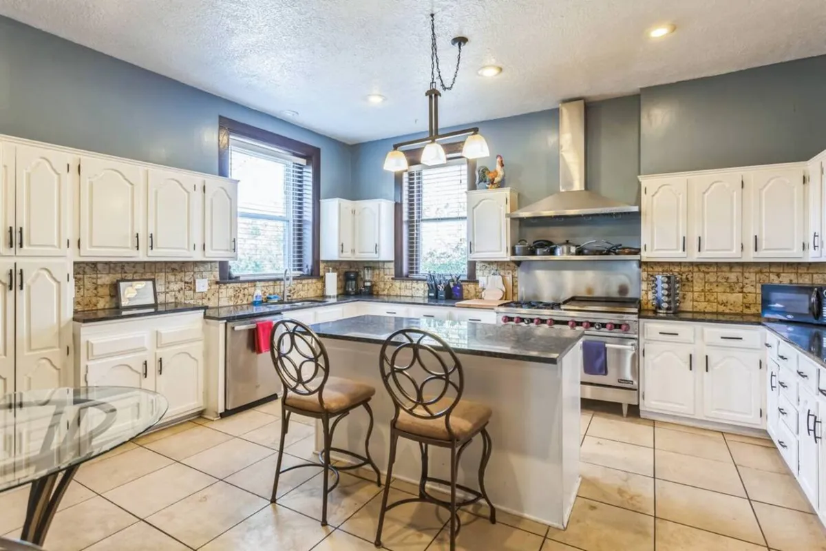 The image features a bright, inviting kitchen with a central island and bar stools. Stainless steel appliances line the space, including a large commercial-grade refrigerator and a professional stove. Generous counter space and natural light from expansive windows create an atmosphere that is both practical and welcoming.