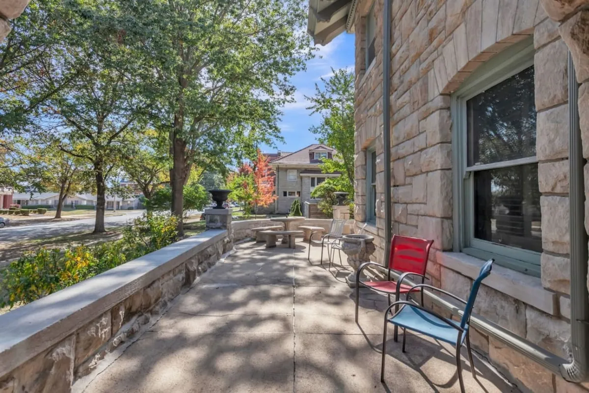 A tranquil outdoor area featuring a stone wall, shaded seating area, lush green trees, and a decorative urn. The space is intimate yet open, ideal for small gatherings, quiet reflection, or appreciating seasonal foliage.