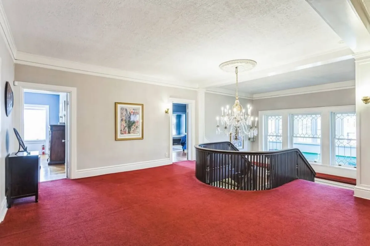 An elegant entry hall with a chandelier, sweeping staircase, wall art, and large windows that fill the space with natural light. The area feels inviting and grand, suggesting a rich historical past and a space where guests gather before venturing out.