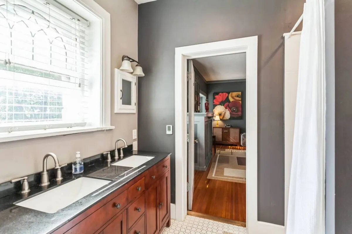 A serene bathroom featuring a dual sink vanity, decorative mirror, and a doorway leading to another room. The design blends modern amenities with period charm, creating a calming space for personal care and quick morning routines.