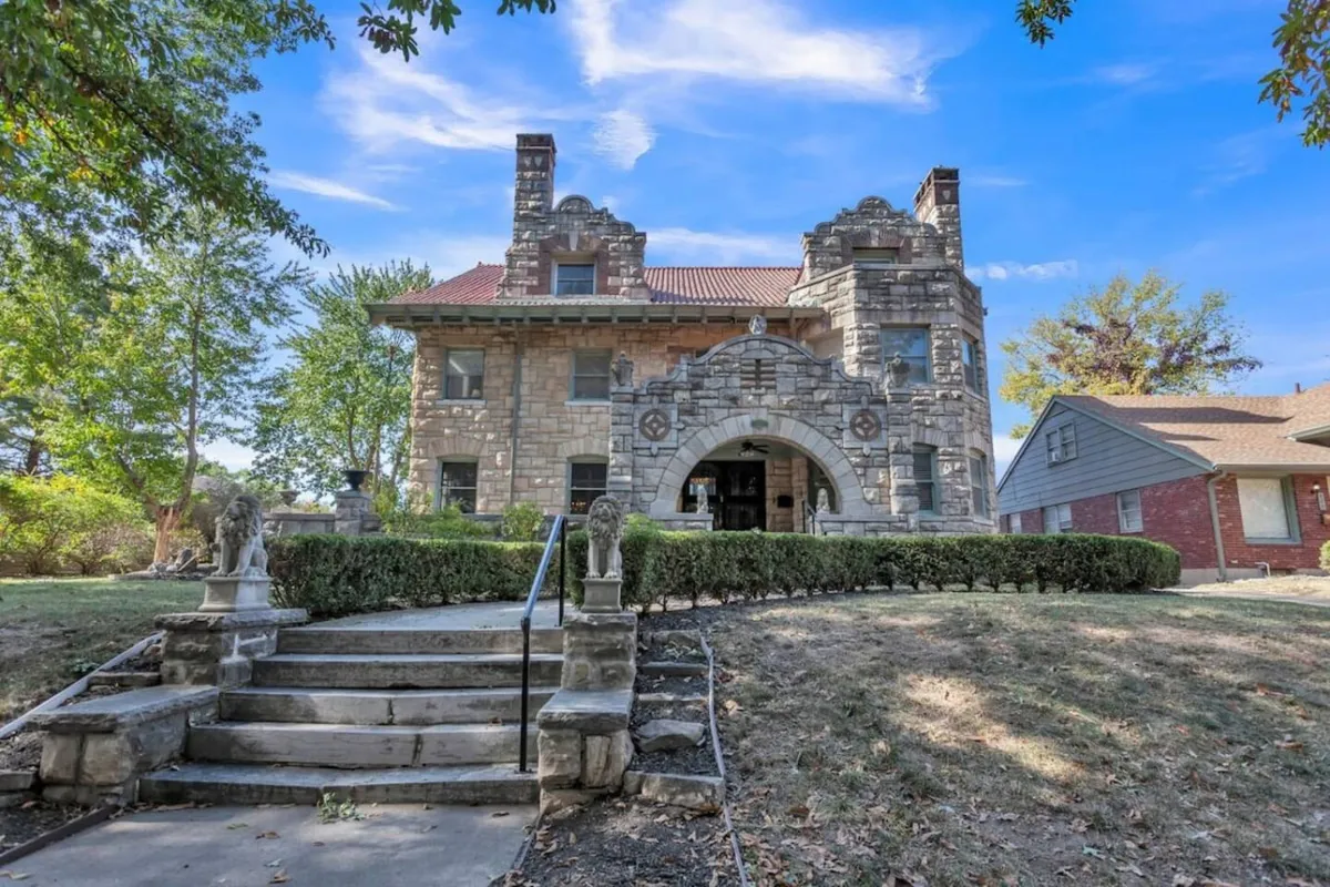 A grand exterior view of the mansion, showcasing ornate stonework, an impressive arched entrance, and a beautifully landscaped front yard. The scene hints at the building's historical significance and its role as a setting for community or cultural events.