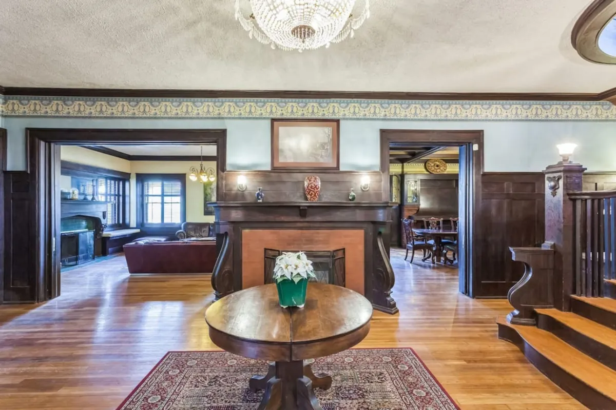 A warmly lit interior featuring a grand chandelier, a classic fireplace, a round table set for gatherings, and an ornate staircase. The space is both elegant and inviting, ideal for intimate discussions or group activities.