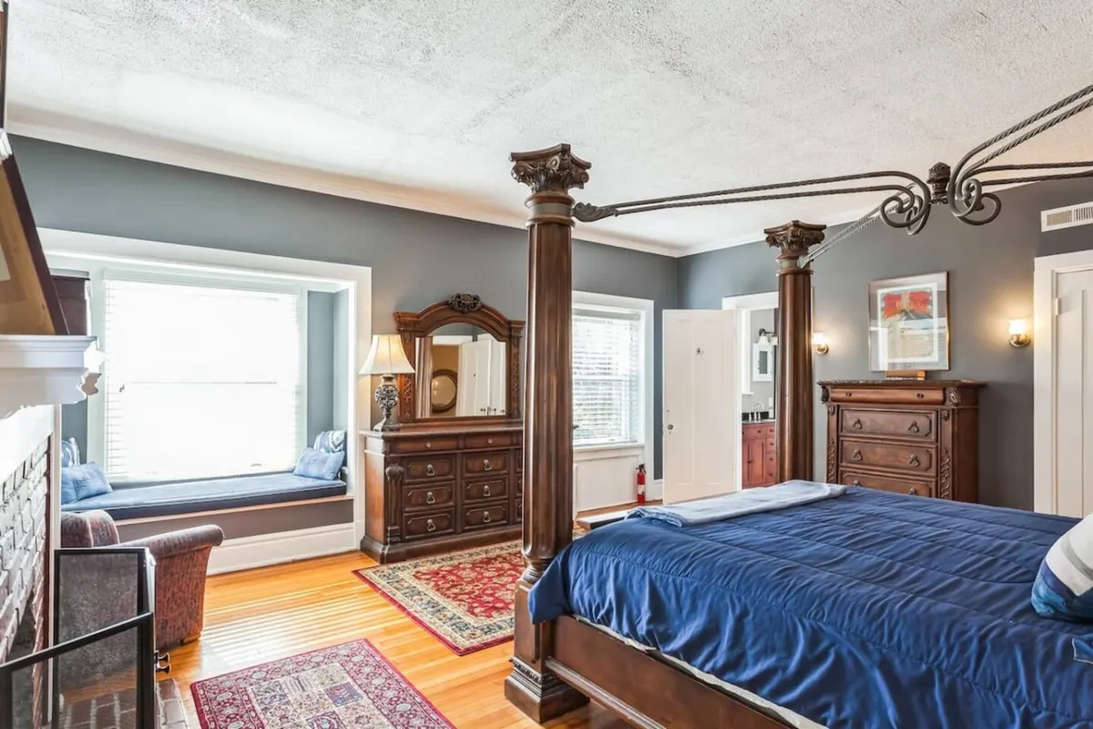 This bedroom features a four-poster bed, a dressing table, cozy window seating, and a soft area rug. The room is bathed in natural light, balancing elegance with comfort, and provides a sense of privacy and tranquility.