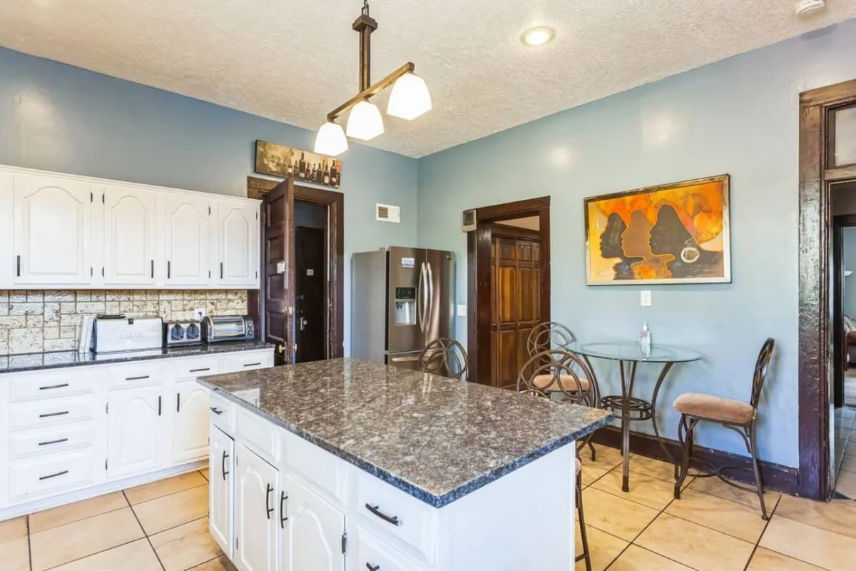 A spacious, modern kitchen featuring a granite island countertop, white cabinetry, stainless steel refrigerator, and wall art, arranged to promote social interaction and culinary creativity.